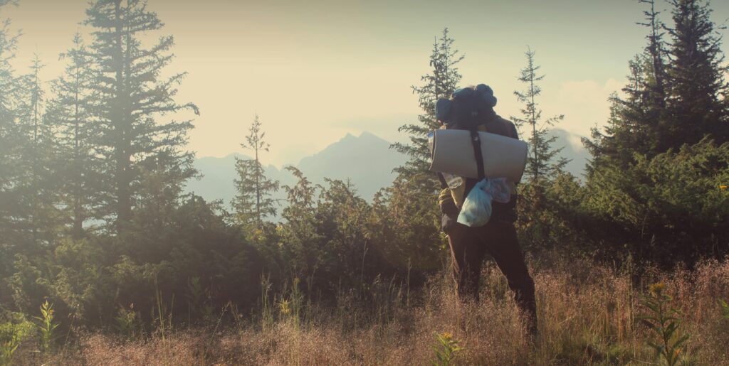 A lone hiker with a backpack and sleeping mat in a sunny mountain landscape.